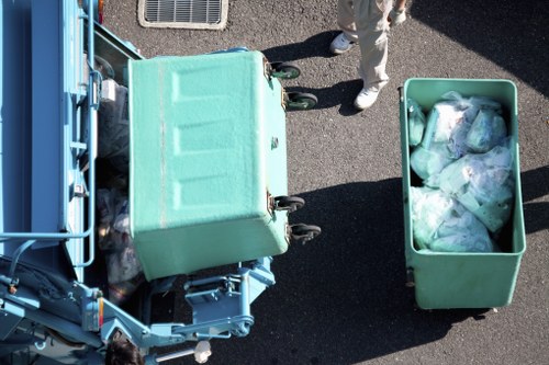 An overhead view of a light green wheeled waste collection bin and a matching larger bin filled with assorted plastic bags, situated on a dark asphalt surface in a parking area. The larger bin is positioned to the right of the smaller bin, which is partially open with some waste visible. In the background, part of a blue waste collection vehicle is visible on the left side, with a person standing nearby, dressed in light-colored clothing and white shoes. The scene suggests a rubbish removal operation, with the bins holding household or commercial waste in preparation for collection, likely indicating a service provided by Commercial Waste Hornsey in the local area.