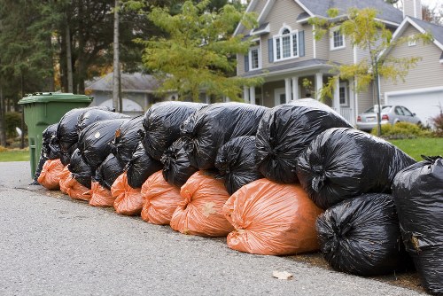 A row of stacked black and orange rubbish bags lined up along a quiet residential street, with black bags on top and orange bags at the bottom. The bags appear to be filled with household or garden waste, with some slightly bulging and tied at the top. Behind the bags, there are green wheelie bins, with a background of a suburban residential area featuring a white and grey detached house with a front garden, a driveway, and parked cars. The street is lined with trees and greenery, suggesting a typical suburban environment, and the scene is captured in daylight with natural lighting. This image relates to waste management services offered by Commercial Waste Hornsey, focusing on rubbish collection and disposal in the Hornsey area, which is part of north London postcode regions.
