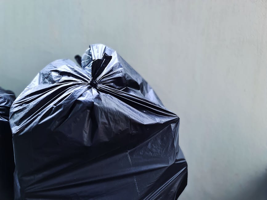 Close-up of a black plastic rubbish bag with a glossy, crinkled surface, tightly sealed and placed against a plain, light-colored wall. The bag's material shows prominent creases and folds, and the to